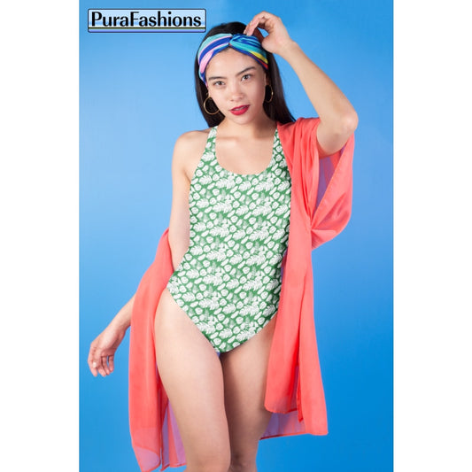 "Tropical allure: A woman stands against a vibrant blue sky, showcasing a one-piece swimsuit from PuraFashions.com adorned with a lush tropical leaves pattern on a serene sea green background, embodying the essence of paradise."