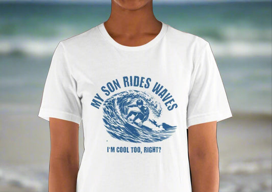 Woman wearing a white T-shirt featuring a design of a guy riding a wave on a surfboard. The text above the design reads "My Son Rides Waves" and below it reads "I'm Cool Too, Right?". Available at PuraFashions.com.
