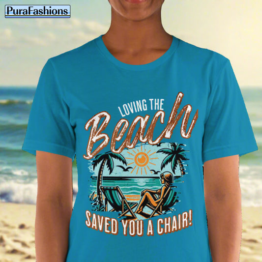 A woman wearing an aqua T-shirt featuring a design of two beach chairs on a tropical beach, with a woman sitting in one chair. Above the design is the text "Loving the Beach" and below it is the text "Saved You A Chair!". Available at PuraFashions.com.