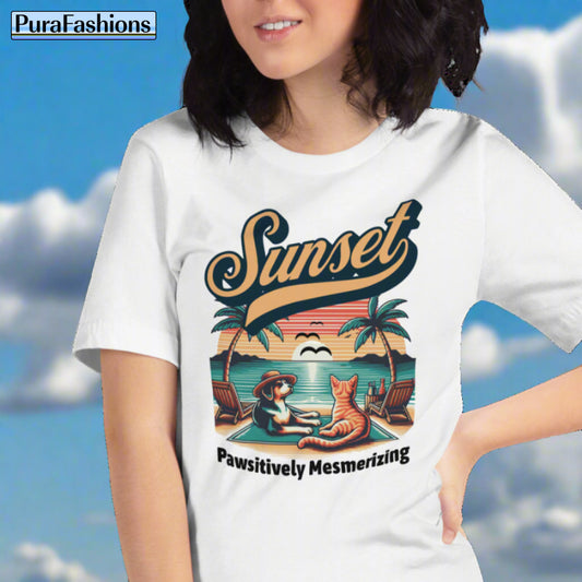 A woman wearing a white T-shirt featuring a design of a dog and cat gazing at a beach sunset. The text above the image reads "Sunset" and the text below reads "Pawsitively Mesmerizing". Available at PuraFashions.com.