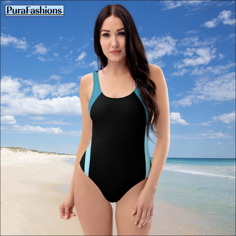 "Beachside elegance: A woman stands gracefully on the sandy shore, showcasing a chic one-piece swimsuit from PuraFashions.com in classic black with stylish blue trim, epitomizing timeless sophistication and coastal charm."